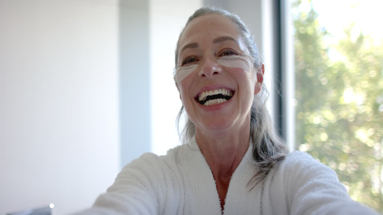 Smiling mature woman in bathrobe with eye patches taking selfie at home