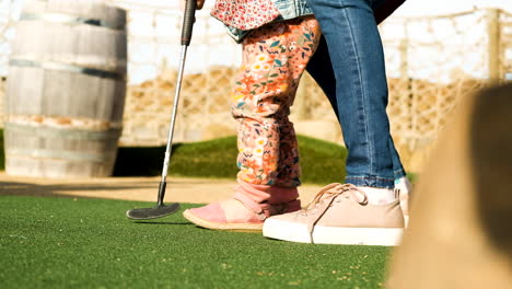 mom assists toddler to make a putt with a pink ball at miniature golf