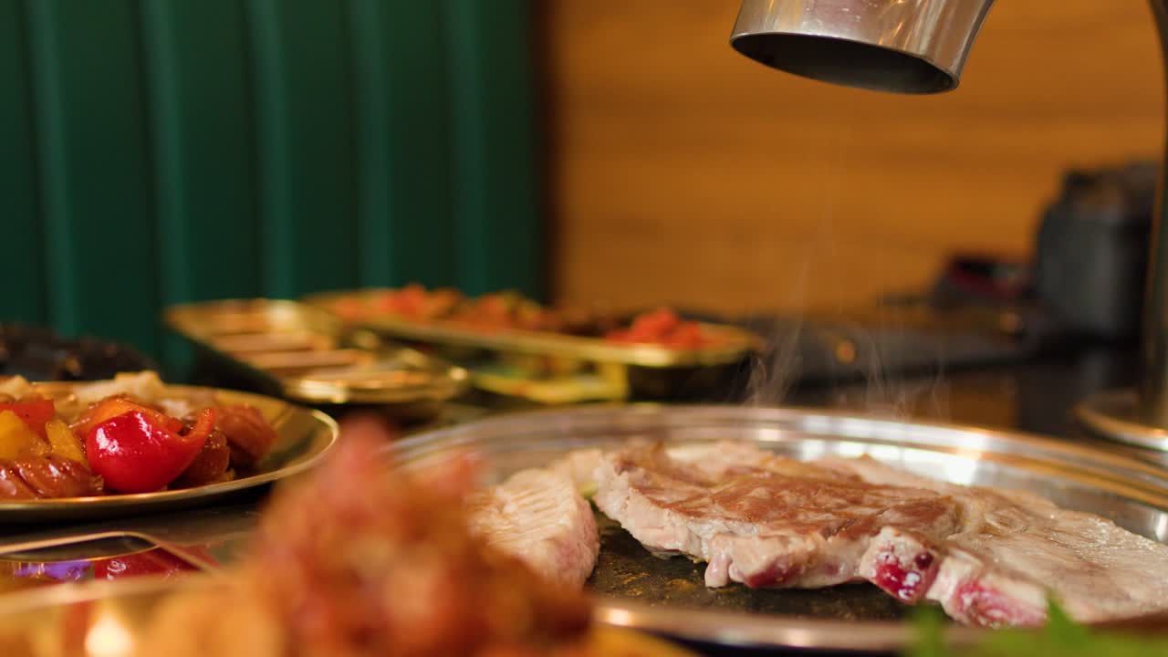 Raw meat and vegetables sizzle on a hot grill pan, steam rising under warm lighting