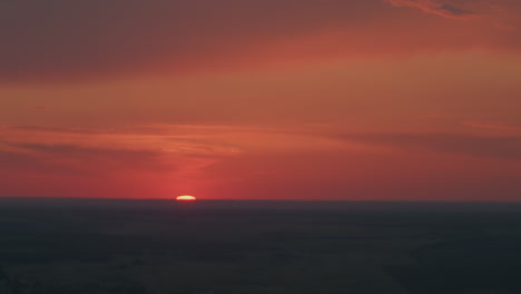 vibrant red orange sunset sinking into distant horizon over flat farmland silhouette under dramatic dusk clouds with subtle lens flare and pastel glow creating serene twilight atmosphere