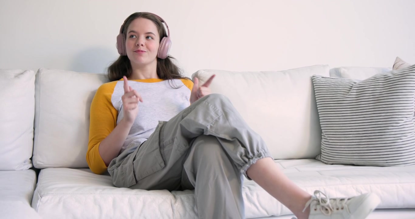 Voksen kvinde sidder på en hvid sofa derhjemme med et lyserødt headset, holder en telefon og tapper i takt til musikken.