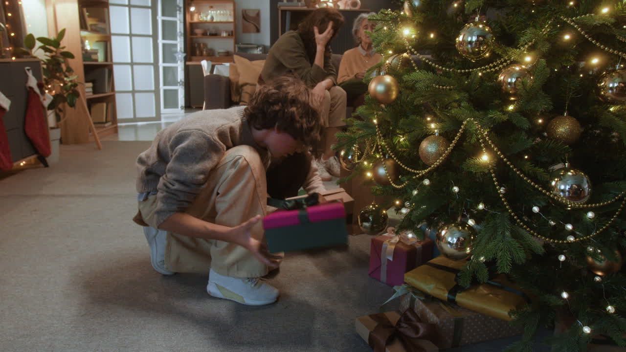 Boy placing presents under Christmas tree with family in background