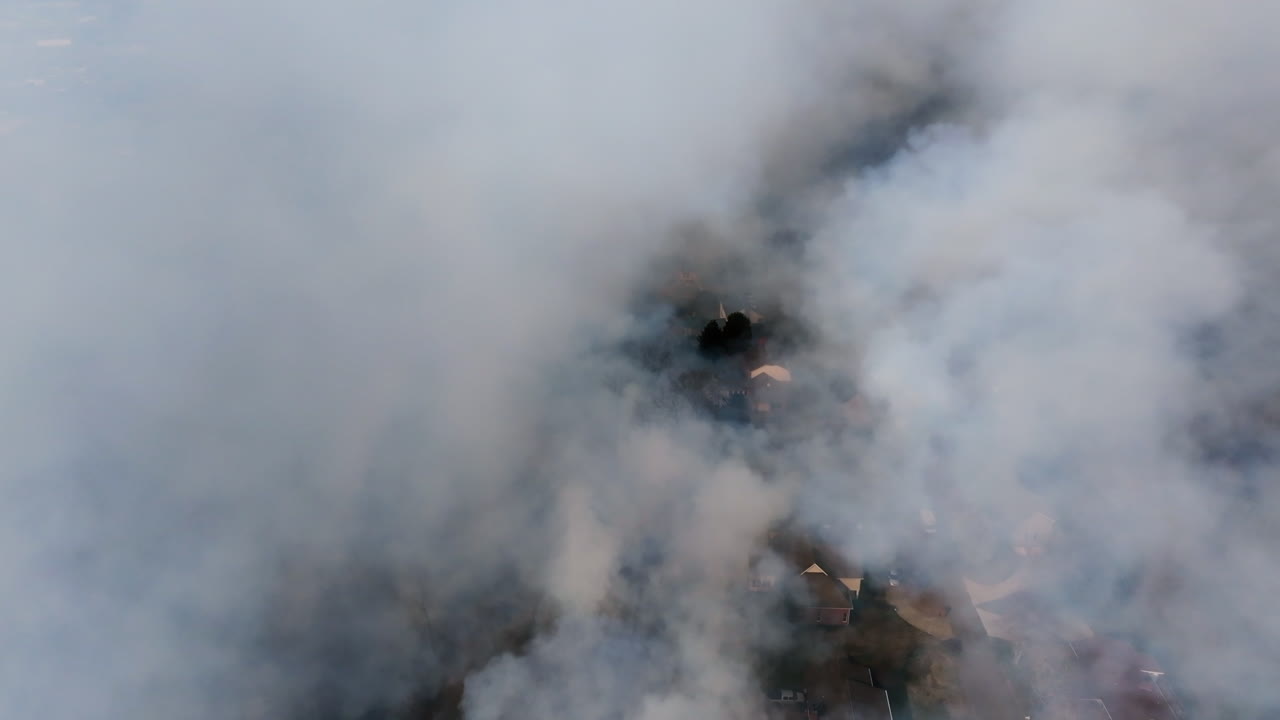 Aerial footage panning around the heavy smoke of a wildfire revealing houses that are very close to the fire.