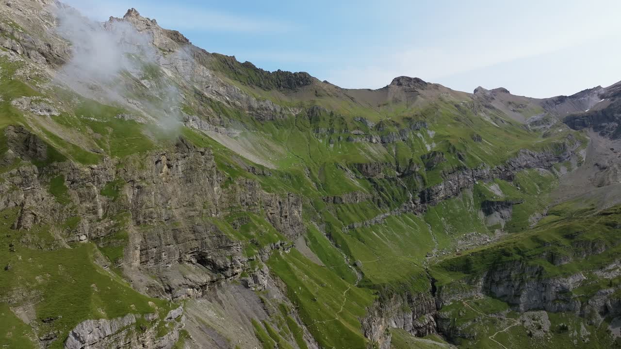 Drone shot of rugged alpine mountains with steep rocky cliffs, layered geological formations, and green meadows scattered across the slopes. Light clouds drift along the ridges