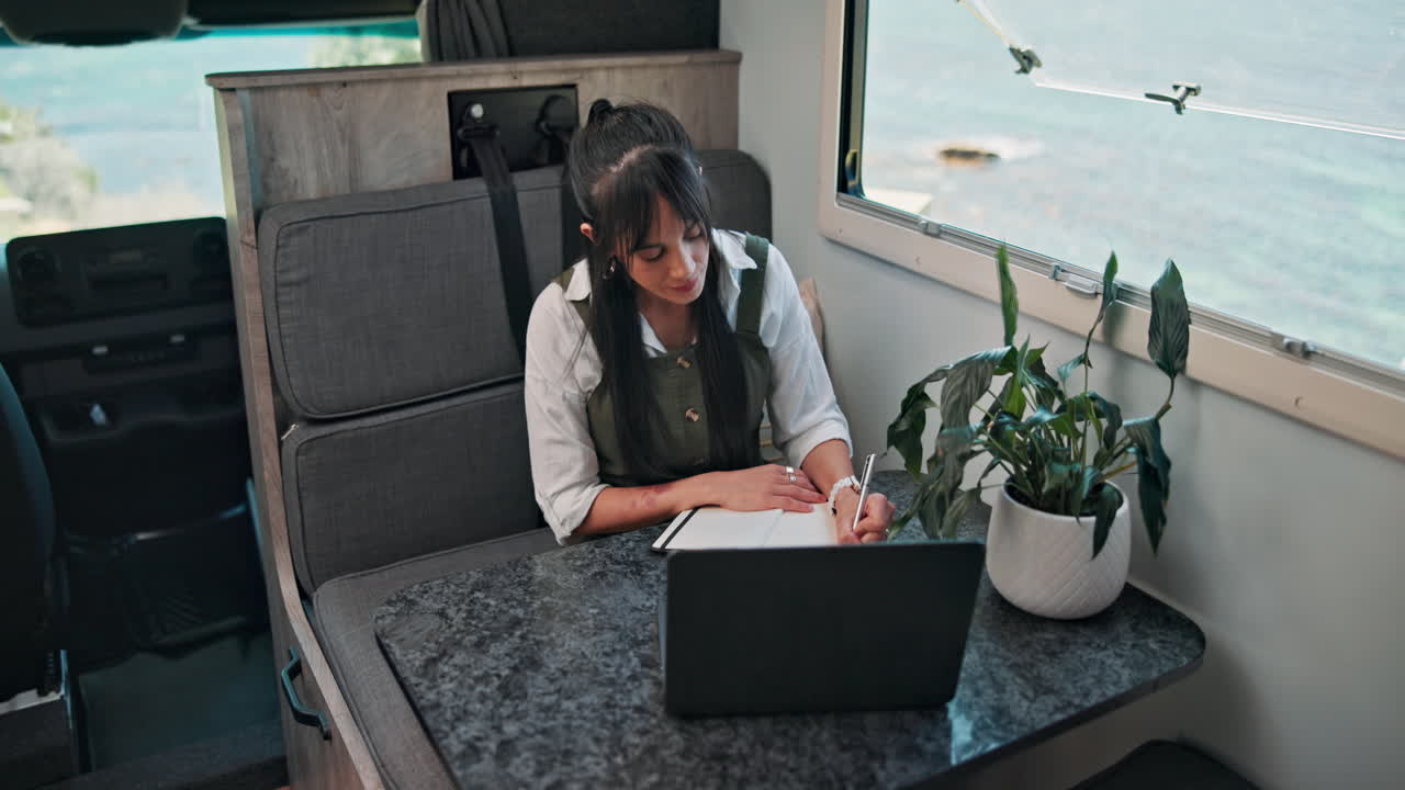 Young Woman Writing in Notebook While Working on Laptop in Camper Van