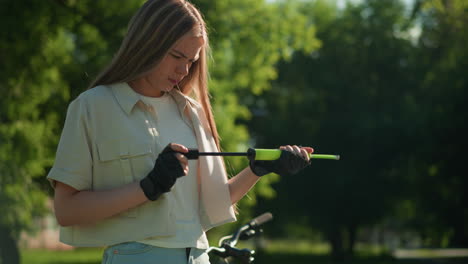 young woman with blonde hair carefully examines green air pump and nozzle in gloved hands, standing outdoors against lush green tree background on a sunny day