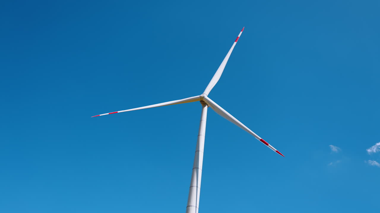 Wind turbine spins under clear blue sky. Tall wind turbine with red and white accents rotates efficiently in the bright sunny sky, harnessing wind energy