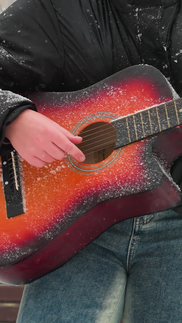 primer plano de manos tocando una vibrante guitarra acústica roja y naranja al aire libre en un parque nevado, la nieve cubre las cuerdas y la manga, mezclando la belleza serena del invierno con la pasión musical