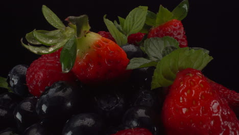 Zooming-out-on-rotating-fresh-forest-berries-in-a-wooden-bowl,-wet-bright-fruits,-strawberries,-blueberries,-raspberries,-4K-macro-shot