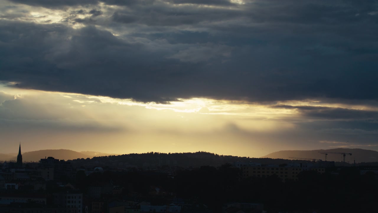 Sun breaks through a gap in heavy sky, casting soft rain shafts over distant Norwegian hills and town silhouettes.