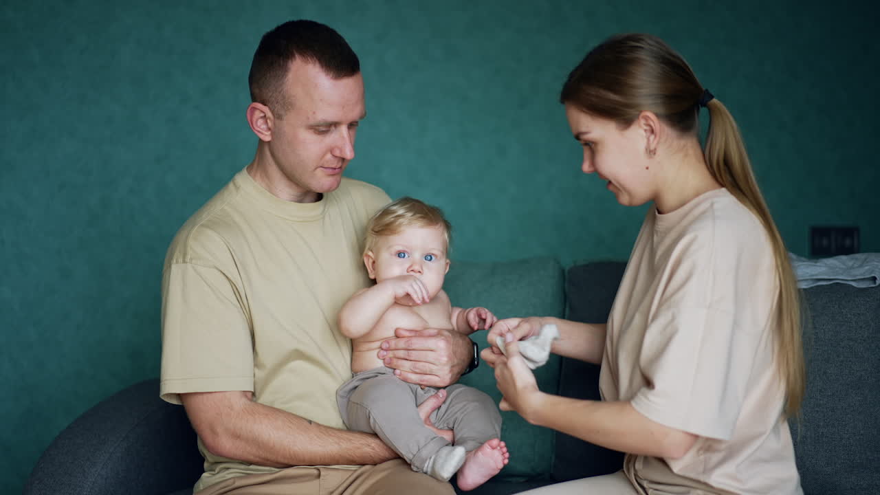 Mom puts on socks on her infant child held by the dad. Parents take care of their cute baby.
