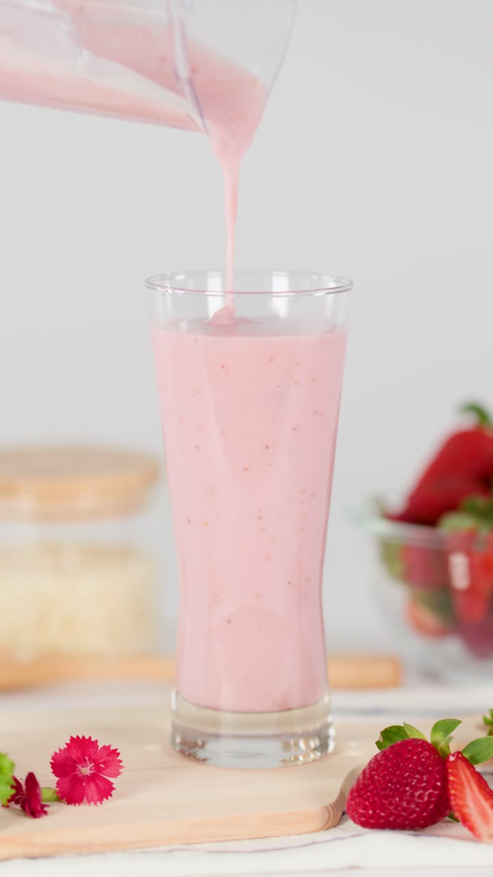 Batido de fresa rosa se vierte suavemente en un vaso, rodeado de fresas frescas, luz diurna brillante, enfoque suave