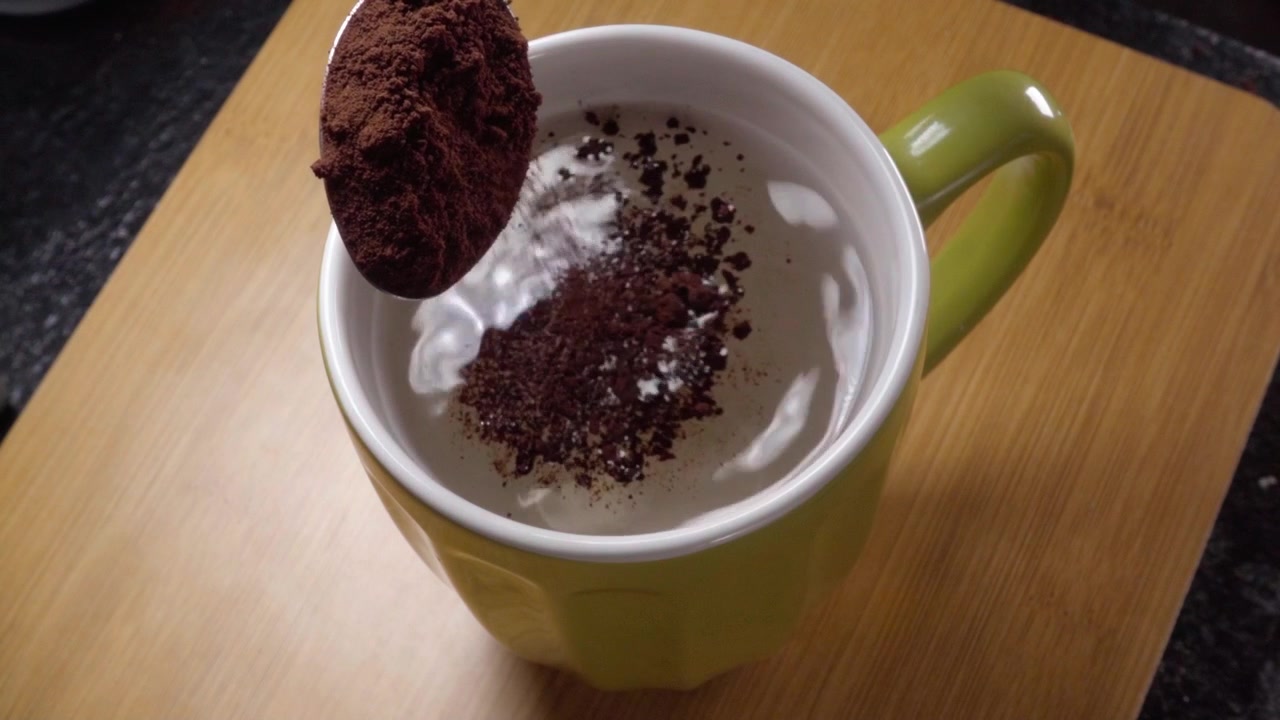 Primo piano di cacao in polvere che cade nel latte all'interno di una tazza di ceramica, creando una ricca bevanda al cioccolato. Processo di miscelazione fluido per la preparazione di bevande fatte in casa su un tavolo di legno