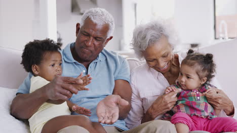 grandparents sitting on sofa at home playing game with baby granddaughters
