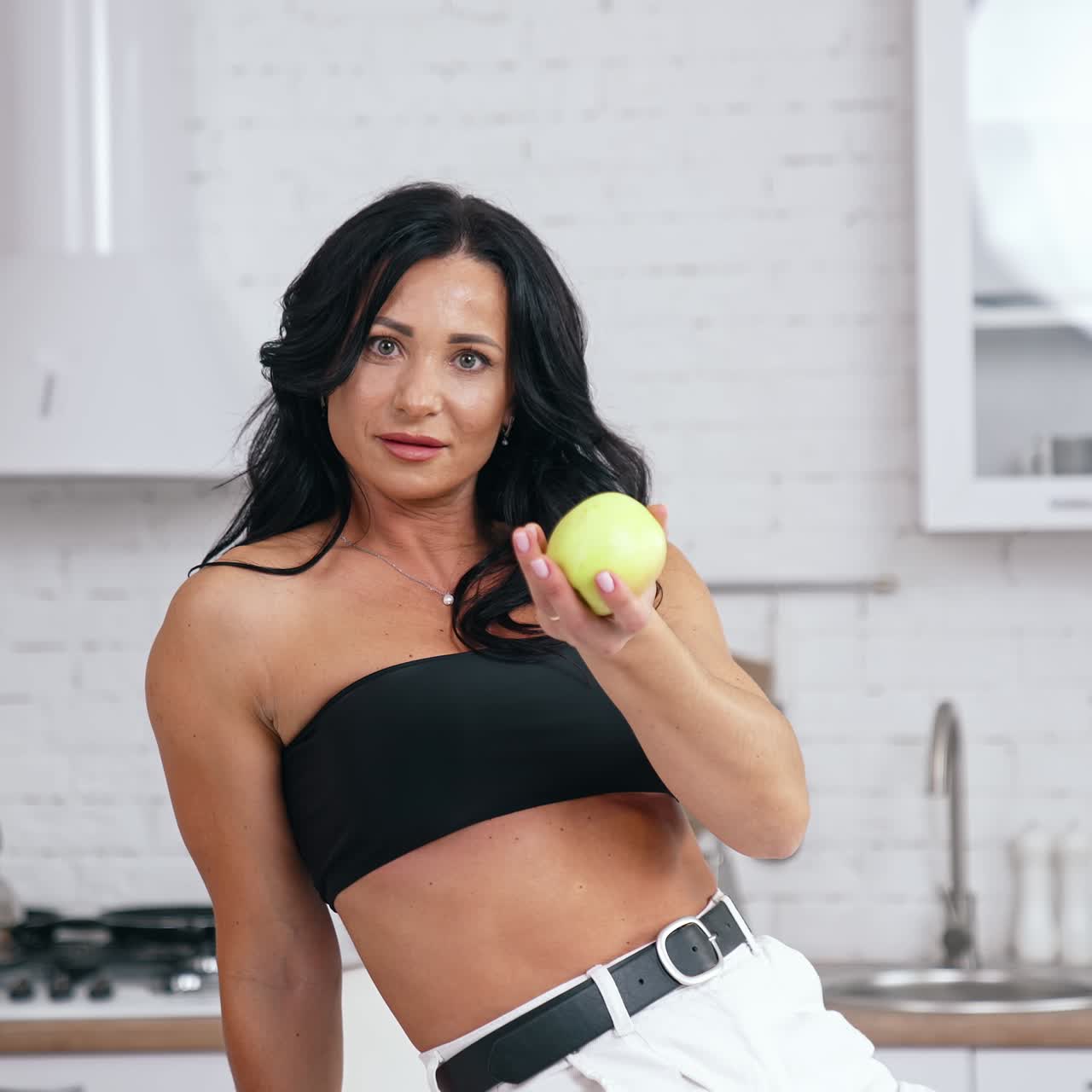 Dark- haired woman with a green apple. Attractive woman sitting on the table in the kitchen and proposing fresh apple. Healthy food