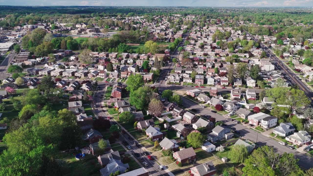 Aerial drone view of a sprawling Philadelphia suburban neighborhood with tree-lined streets, single-family homes, rail lines, and spring foliage throughout.