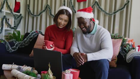 video de una feliz pareja diversa en sombreros de santa haciendo una llamada de video de computadora portátil de navidad en casa