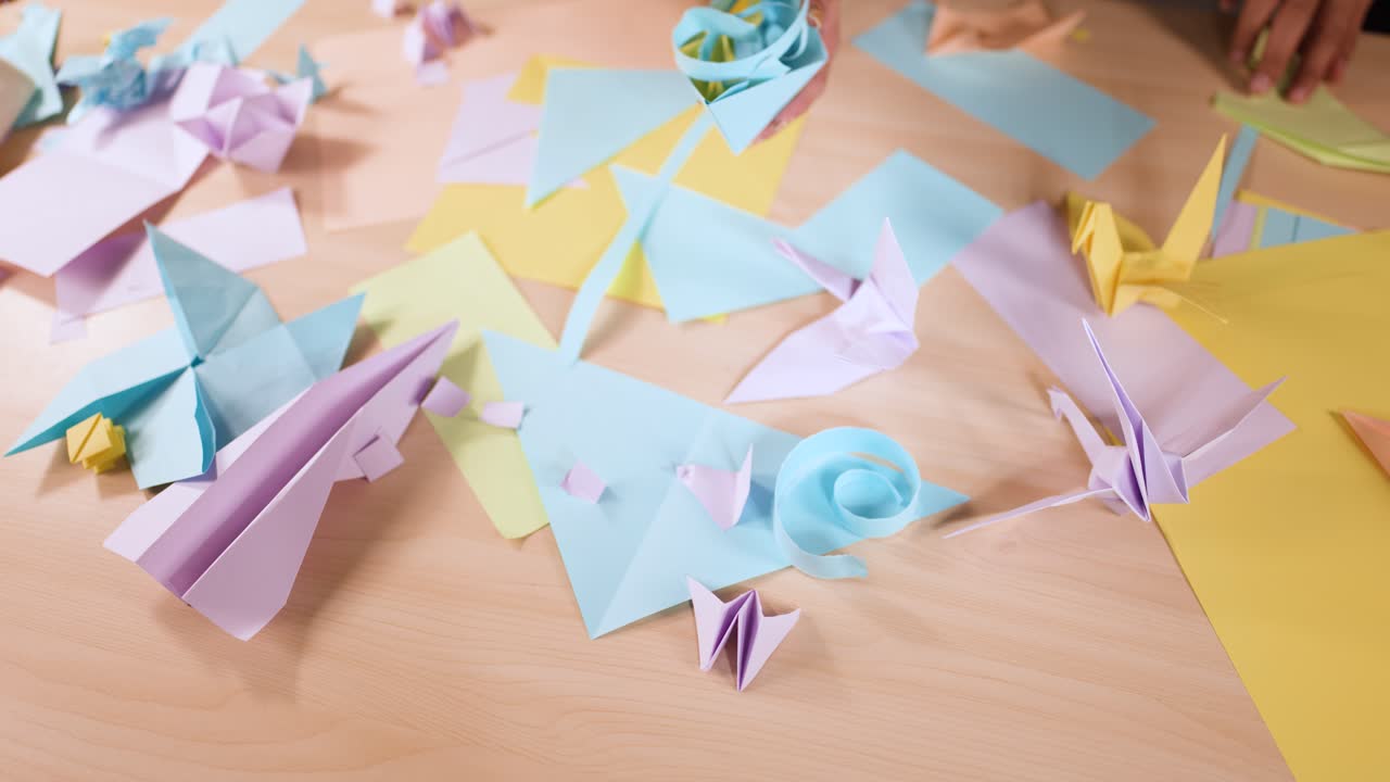 Overhead shot of hands carefully placing various pastel origami paper creations on a wooden surface
