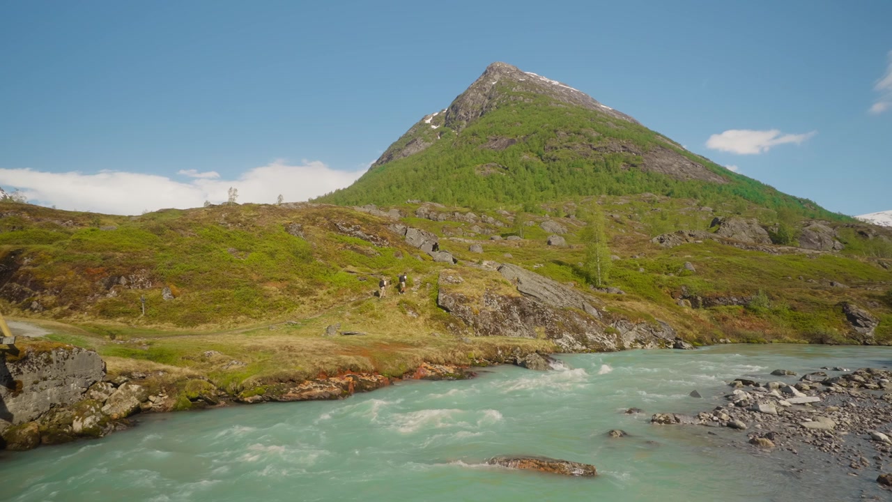Inquadratura statica dal basso di due persone che camminano lungo un fiume glaciale turchese con montagne sullo sfondo sotto un cielo estivo limpido e azzurro in Norvegia. Paesaggio alpino da viaggio suggestivo
