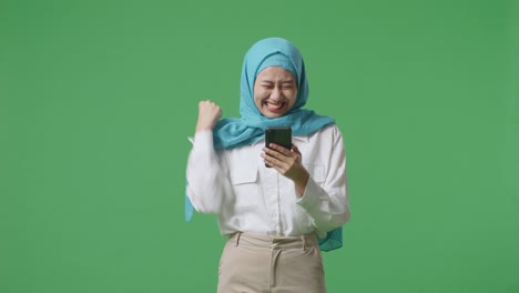 asian muslim woman looking at smartphone then screaming goal celebrating while standing in the green screen background studio