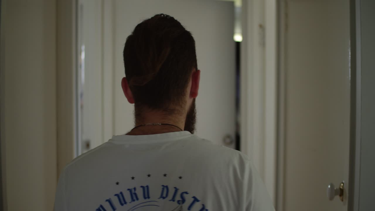 A man with long hair and a beard walks down a narrow, white hallway. He’s wearing a white shirt with blue text printed on the back, he is heading toward a slightly ajar door.