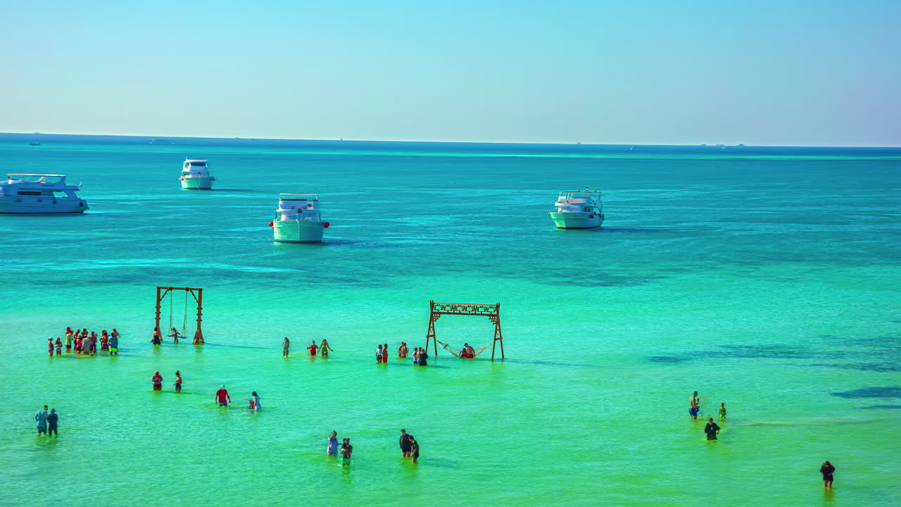 관광객들이 얕은 물에서 장난감을 가지고 놀고 요트가 더 깊은 곳에서 앞뒤로 항해하는 청록색 바다의 만