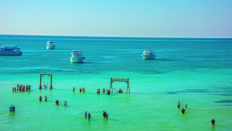 a bay in the ocean with turquoise water where tourists play on toys in the shallow water and yachts sail back and forth in the deeper part