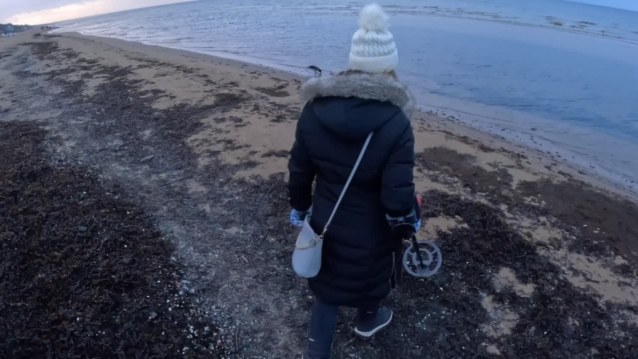 A woman walks along a cold shoreline carrying a metal detector, moving toward the waterline as waves gently reach the beach