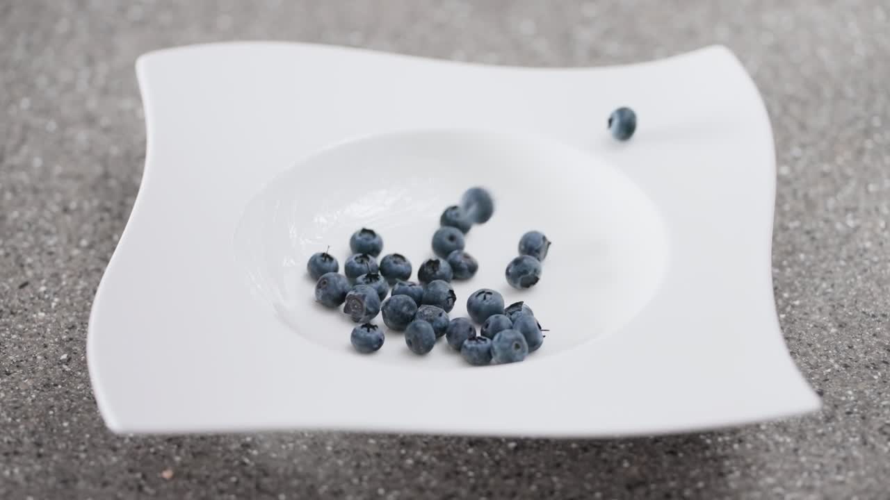 Close-up of ripe, organic blueberries cascading into a stylish white bowl on a grey surface, capturing the dynamic motion of healthy food preparation.