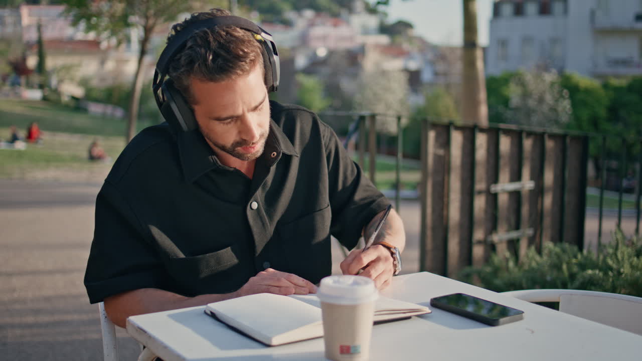Nærbilde av mann med hodetelefoner som skriver i notatbok i en sommerpark. Frilanser lytter til musikk.