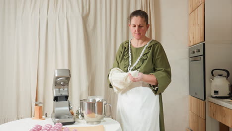 baker in green dress and apron fills piping bag with batter or frosting, preparing for decorative baking process in cozy home kitchen setup with mixer, metal bowl, and piping tools on table