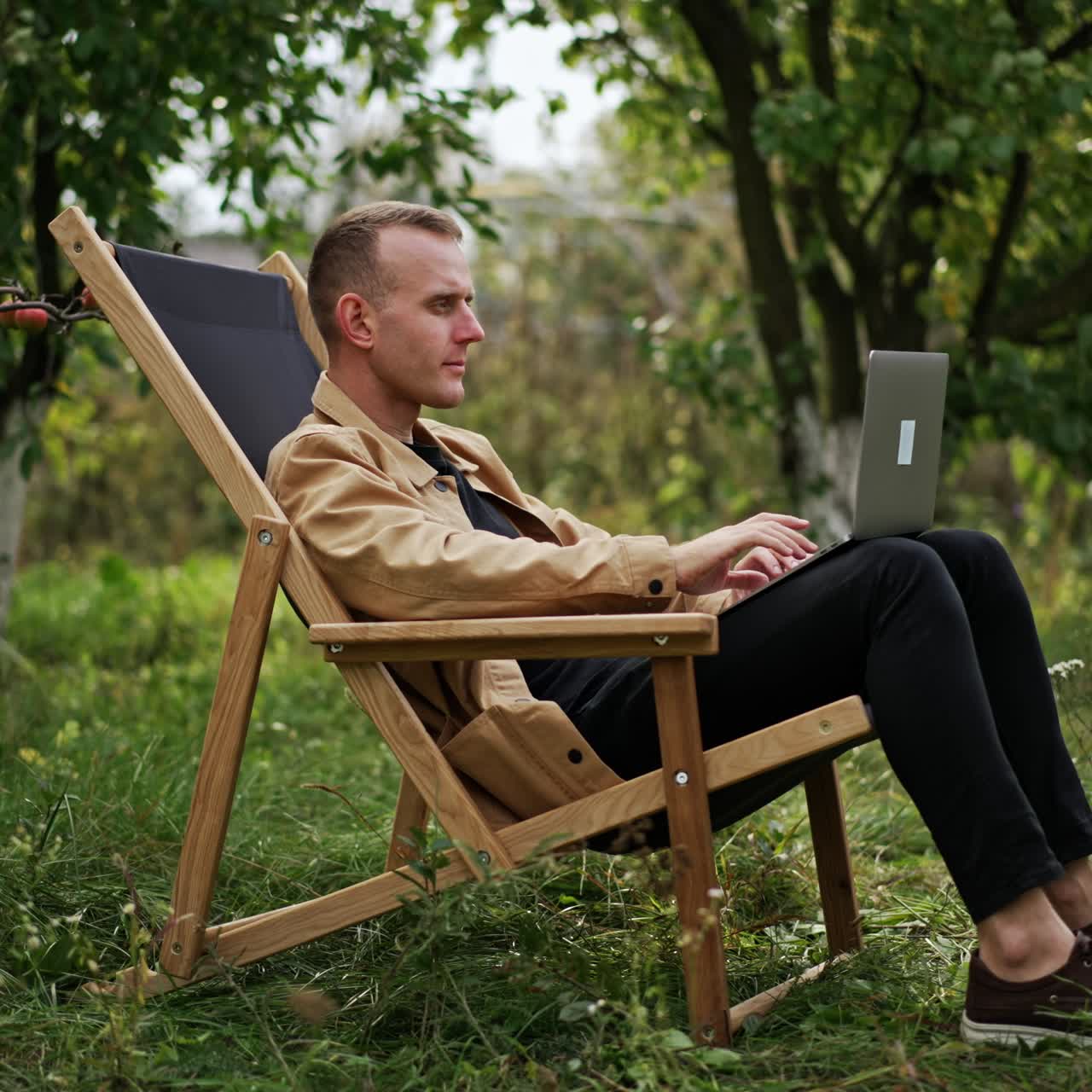 Busy man focused on his work on laptop sitting in garden. Camera slowly approaching freelancer who finishes work, closes laptop and leans back in his chair
