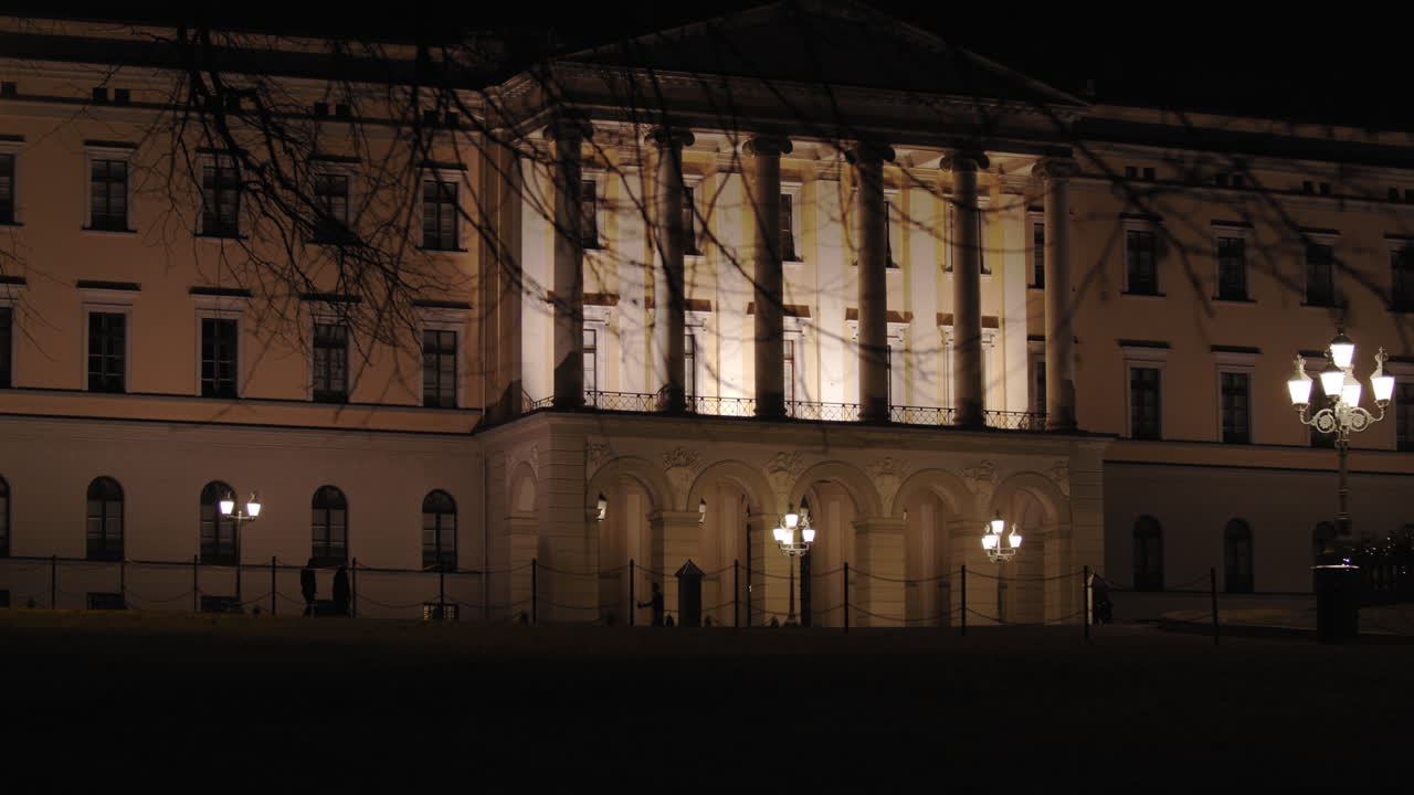 Slow motion close-up to wide 4K shot with parallax motion of tree revealing front of the illuminated Norwegian Royal Palace on top of Karl Johan street, at night in Oslo Norway