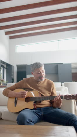vertical video of senior biracial man sitting playing guitar and singing at home, slow motion