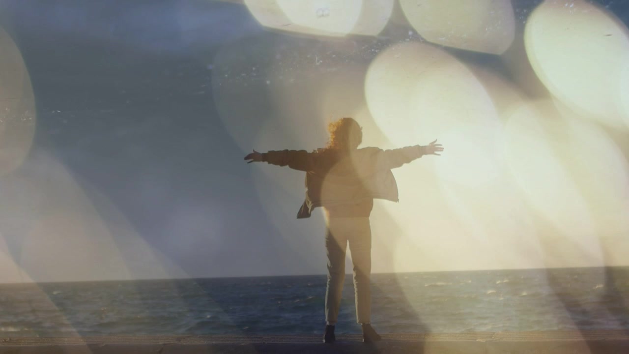 Woman approaching sea wall, straightening and spreading arms as bokeh shifting, creating promo shot