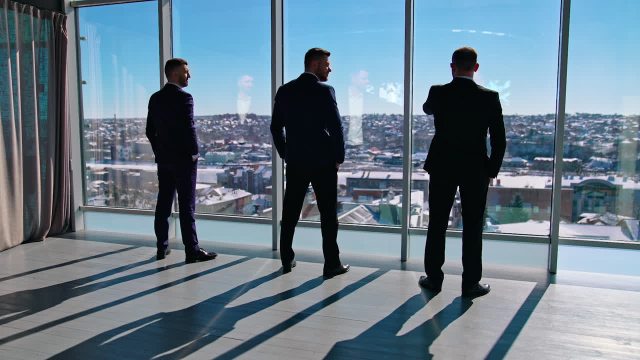 Serious businessmen standing near the windows. Elegant entrepreneurs in suits listen to their partner indoor the skyscraper. Back view.