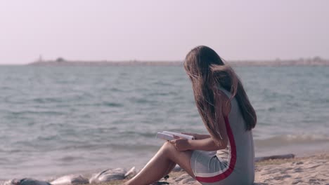 young woman with long hair sits on beach and reads book