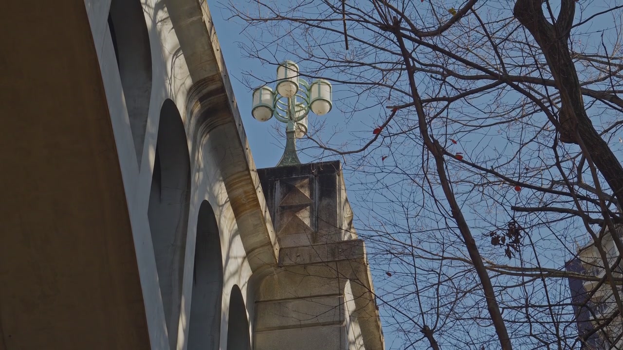 L'arco in pietra del Ponte Otonashi e un lampione vintage ornato si stagliano contro un cielo azzurro pallido, incorniciati da rami invernali spogli, ripresi con un'inquadratura statica dal basso verso l'alto alla luce naturale del giorno
