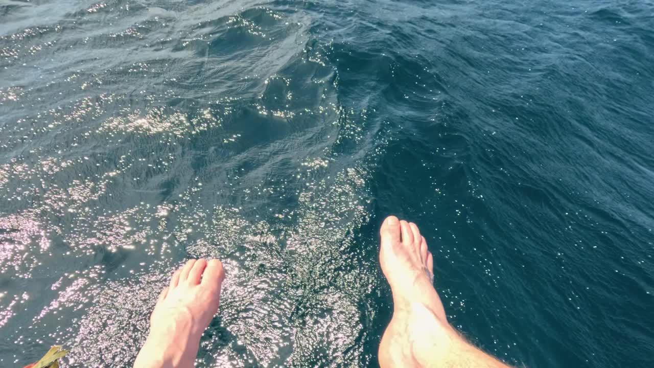 Bare feet dangle above sunlit ocean waves, gentle movement, natural daylight, relaxed vacation atmosphere