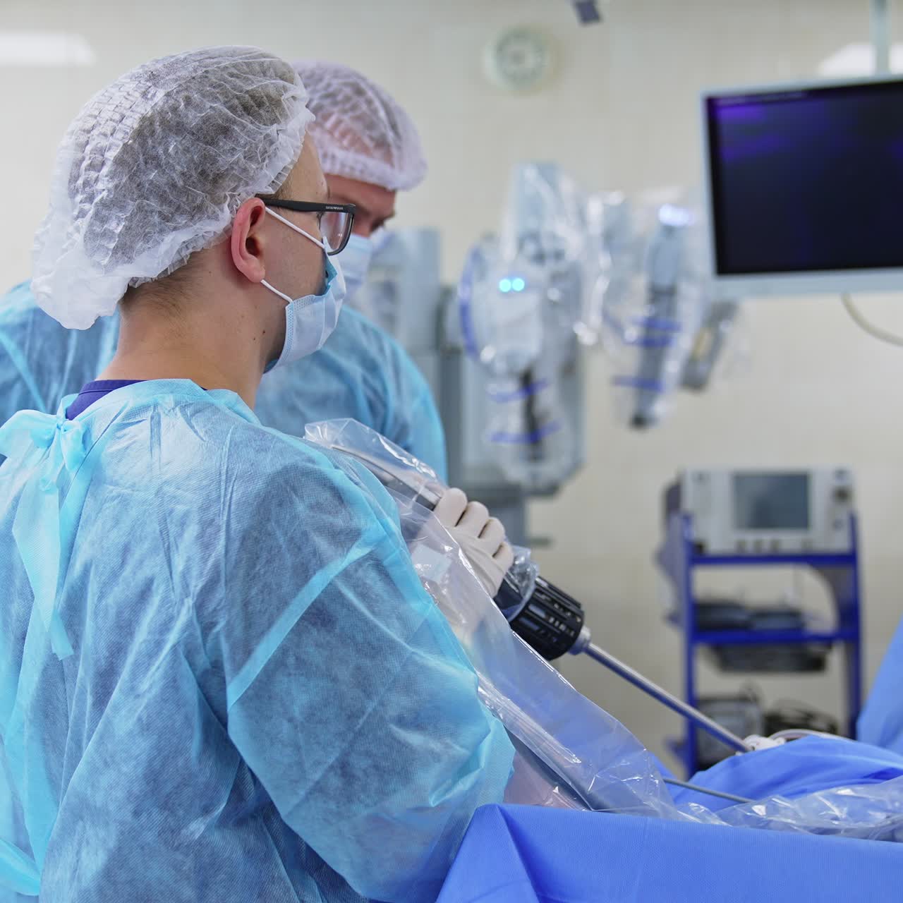 Younger male surgeon holds long device attached to a patient. Chief doctor stands beside. Highly-equipped operational theatre at backdrop in blur
