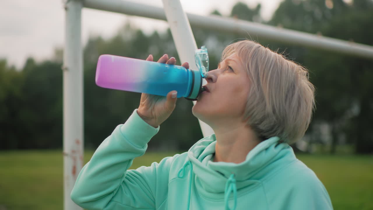 Elegant woman standing on school field, resting against goalpost while sipping coffee from vibrant bottle, casually looking around peaceful green surroundings with relaxed posture