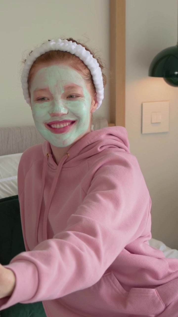 Video verticale: Donna sorridente con felpa rosa, fascia per capelli e maschera verde che reagisce a un amico sul letto