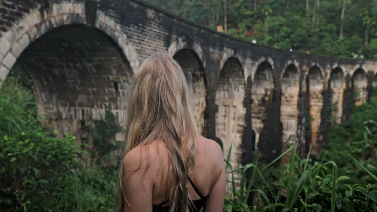 A captivating scene in Ella, Sri Lanka, where a woman walks through the lush jungle, gazing towards the iconic Nine Arches Bridge.