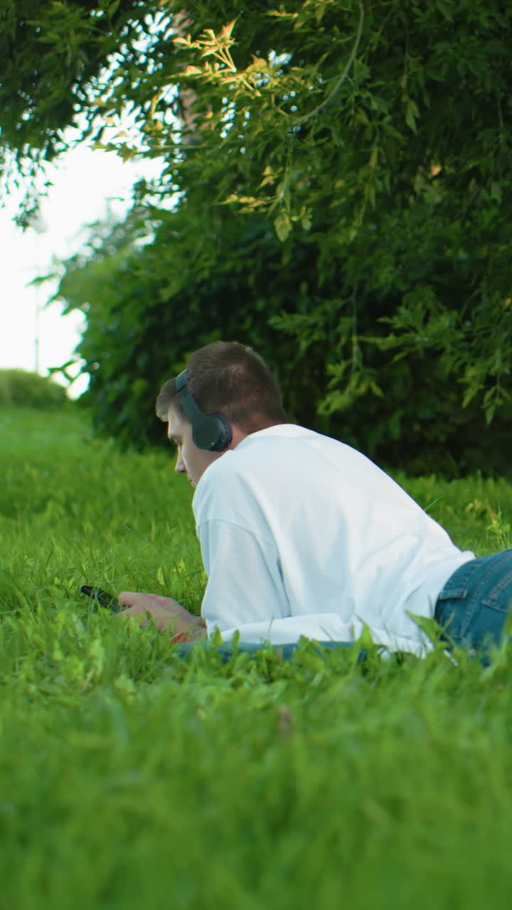 잔디에 누워있는 남자가 헤드폰과 청바지를 입고 청바지에 집중하고, 활기찬 초록색과 나무로 둘러싸여 있으며, 멀리 떨어진 배경에서 고 있는 사람들이 있습니다.