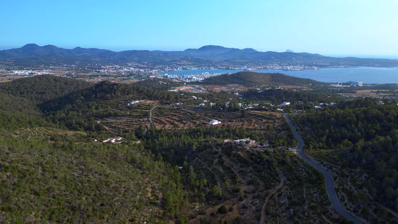 Road winding through a vibrant green forest in Ibiza, Spain, leading down towards the beautiful San Antonio Bay. Magic aerial view flight ascending drone