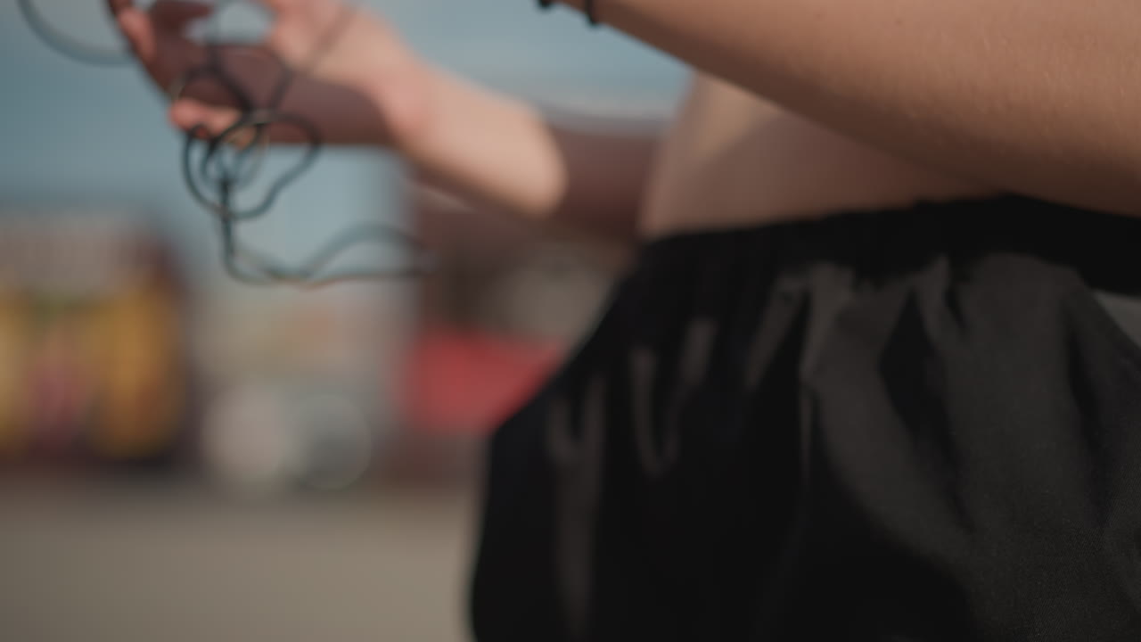 Ragazza che regola il telefono e le cuffie all'aperto, top corto bianco e pantaloncini neri, polsini ai polsi, sfondo di strada illuminata dal sole con segnaletica sfocata, mani in primo piano che collegano il cavo e scorrono