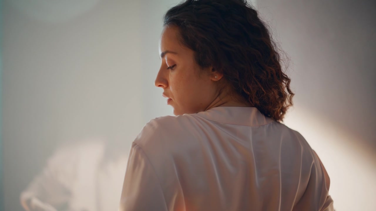 Donna con capelli ricci che si tocca le ciocche, vista da dietro. Primo piano di una donna sognante nella luce del mattino.
