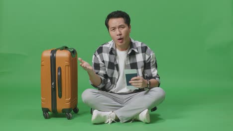 full body of wondering asian male traveler with luggage holding passport and sitting doubtfully in the green screen background studio