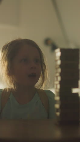 young woman starts game by pulling wooden block from tower and explaining rules to children. family spends game night sitting at small cozy table