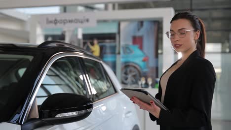 a woman standing by a vehicle inspecting a tablet screen. a woman is standing next to a vehicle, looking at a tablet. she is gesturing towards the automotive design of the car while admiring the automotive exterior and automotive mirror